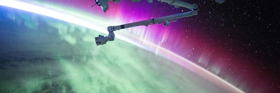 View of colorful aurora over Earth's atmosphere seen from space with part of a spacecraft arm visible.